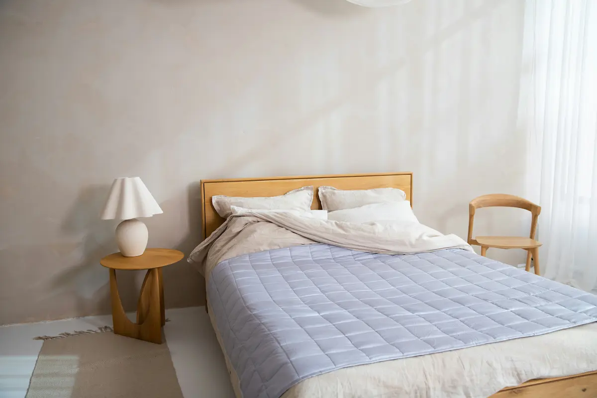 Minimalist bedroom with light wood bed frame, beige bedding and light blue comforter, wooden side table and white lamp